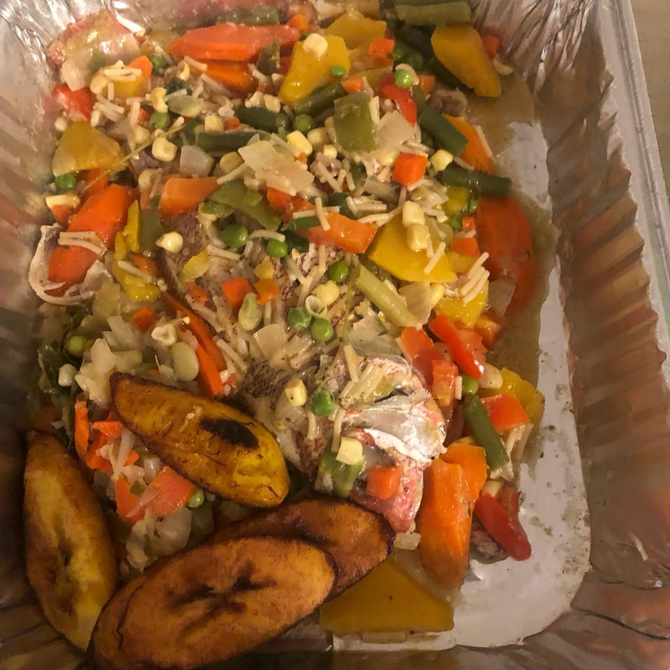 Foil tray of seasoned fish with mixed vegetables, rice, corn, peas, peppers, and fried plantains.