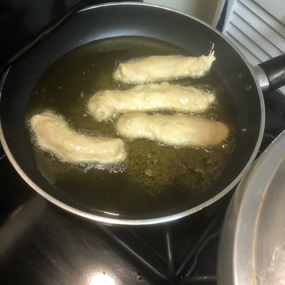 Four battered food pieces fry in bubbling oil inside a black skillet on a stovetop.