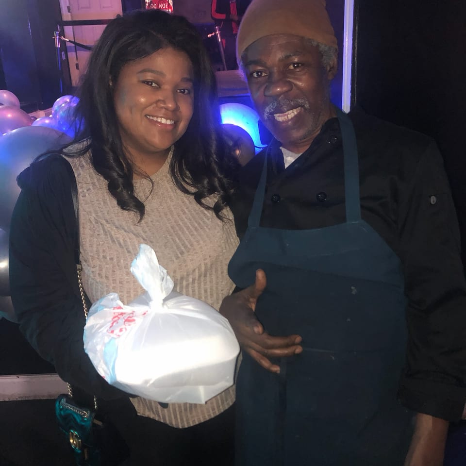 Smiling woman and man pose together in a dimly lit venue with blue lighting and balloons, holding a white takeout container.