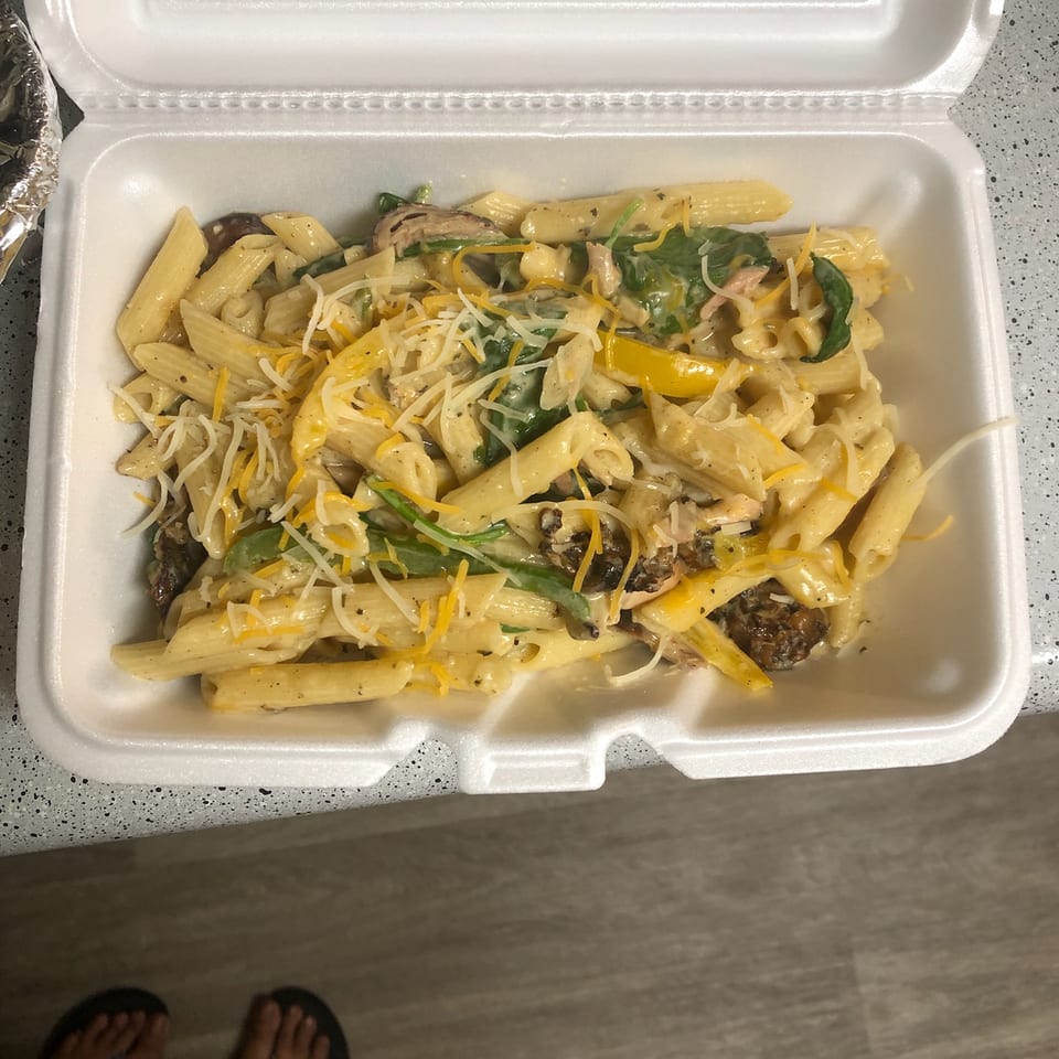 Open takeout container holding creamy penne pasta with chicken, mushrooms, spinach, peppers, and shredded cheese.